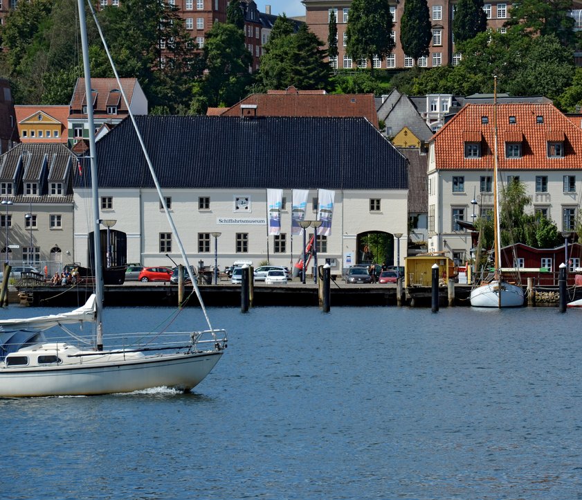 Auf dem Foto ist das Hauptgebäude des Flensburger Schifffahrtsmuseums abgebildet. Das Foto ist vom Ostufer aus aufgenommen, sodass vor dem Museum die Förde zu sehen ist. Von links kommt ein modernes Segelschiff ins Bild reingesegelt. 
