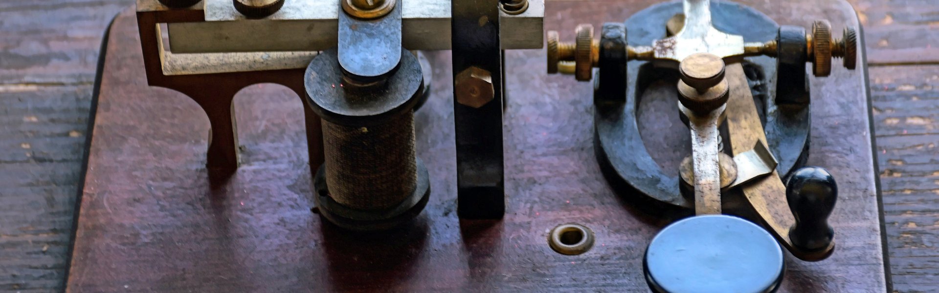 Das Foto zeigt einen alten Morse-Apparat. Zu sehen sind Spulen und ein Taster. Sie sind auf einem dunklen Stück Holz montiert. Der Apparat steht auf einer alten Tisch-Platte. 