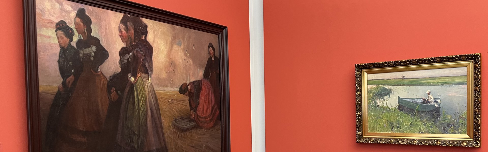 Das Foto zeigt zwei Gemälde in einem Ausstellungsraum. Die Bilder hängen vor einer roten Wand. Auf dem rechten Gemälde sind sechs Frauen abgebildet. Alle tragen Tracht. In der rechten Bildhälfte befinden sich vier Frauen. Sie stehen als Gruppe zusammen. Links etwas weiter hinten hocken zwei Frauen. Auf dem anderen Gemälde ist ein Mann in einem kleinen Ruderboot abgebildet. Er hat eine Angel in der Hand.