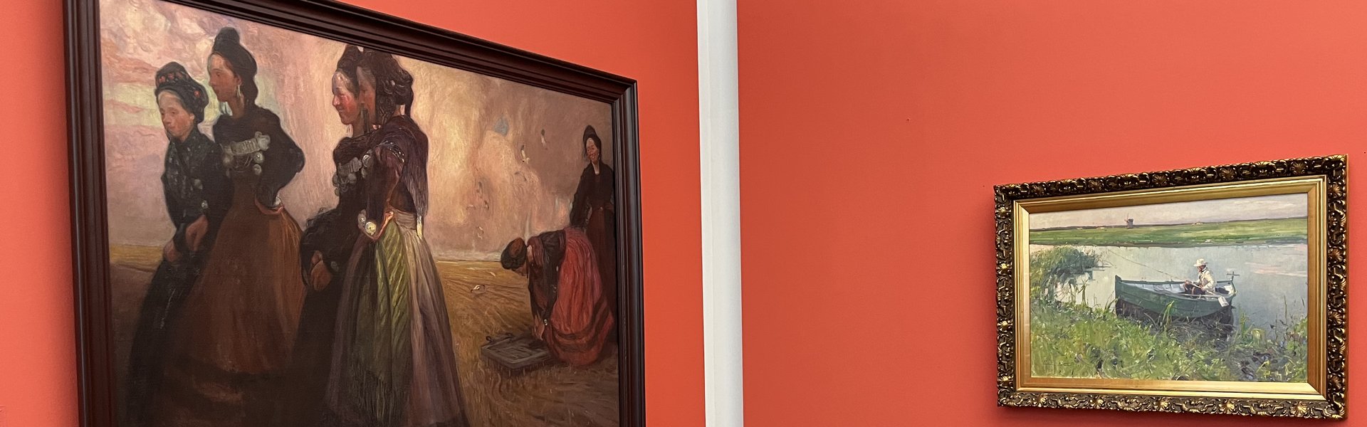 Das Foto zeigt zwei Gemälde in einem Ausstellungsraum. Die Bilder hängen vor einer roten Wand. Auf dem rechten Gemälde sind sechs Frauen zu sehen. Alle tragen Tracht. Eine Tracht ist eine besondere Bekleidung, die oft in bestimmten Regionen oder zu bestimmten Anlässen getragen wird. Auf der rechten Seite des Bildes stehen vier Frauen zusammen in einer Gruppe. Links, etwas weiter hinten, hocken zwei Frauen. Auf dem anderen Gemälde ist ein Mann in einem kleinen Ruderboot zu sehen. Er hält eine Angel in der Hand.