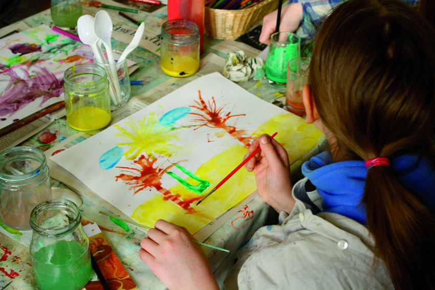 Ein Kind malt mit verschiedenen Farben und Pinseln ein Bild. Das Bild zeigt Palmen am Strand. Auf dem Tisch stehen Farbtöpfe und weitere Malutensilien.